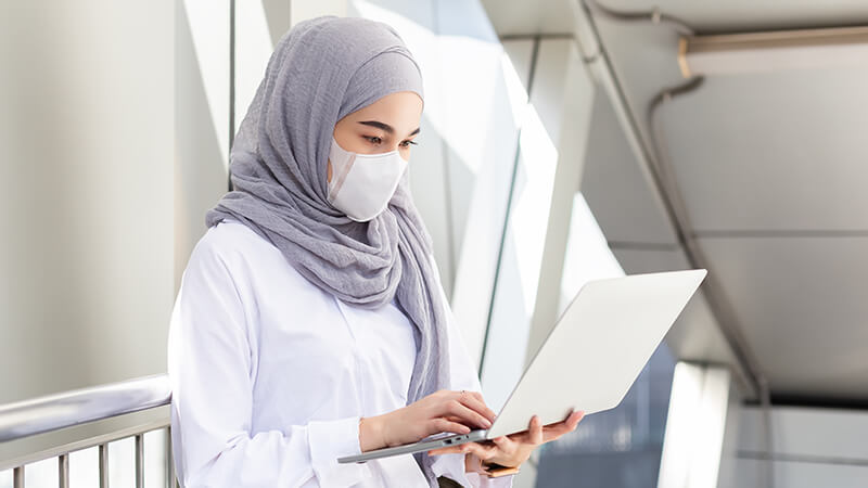 woman wearing a grey hijab and mask using a laptop