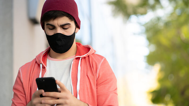man wearing a tight fitting beanie in a hoodie and wearing a mask looking down at a mobile phone